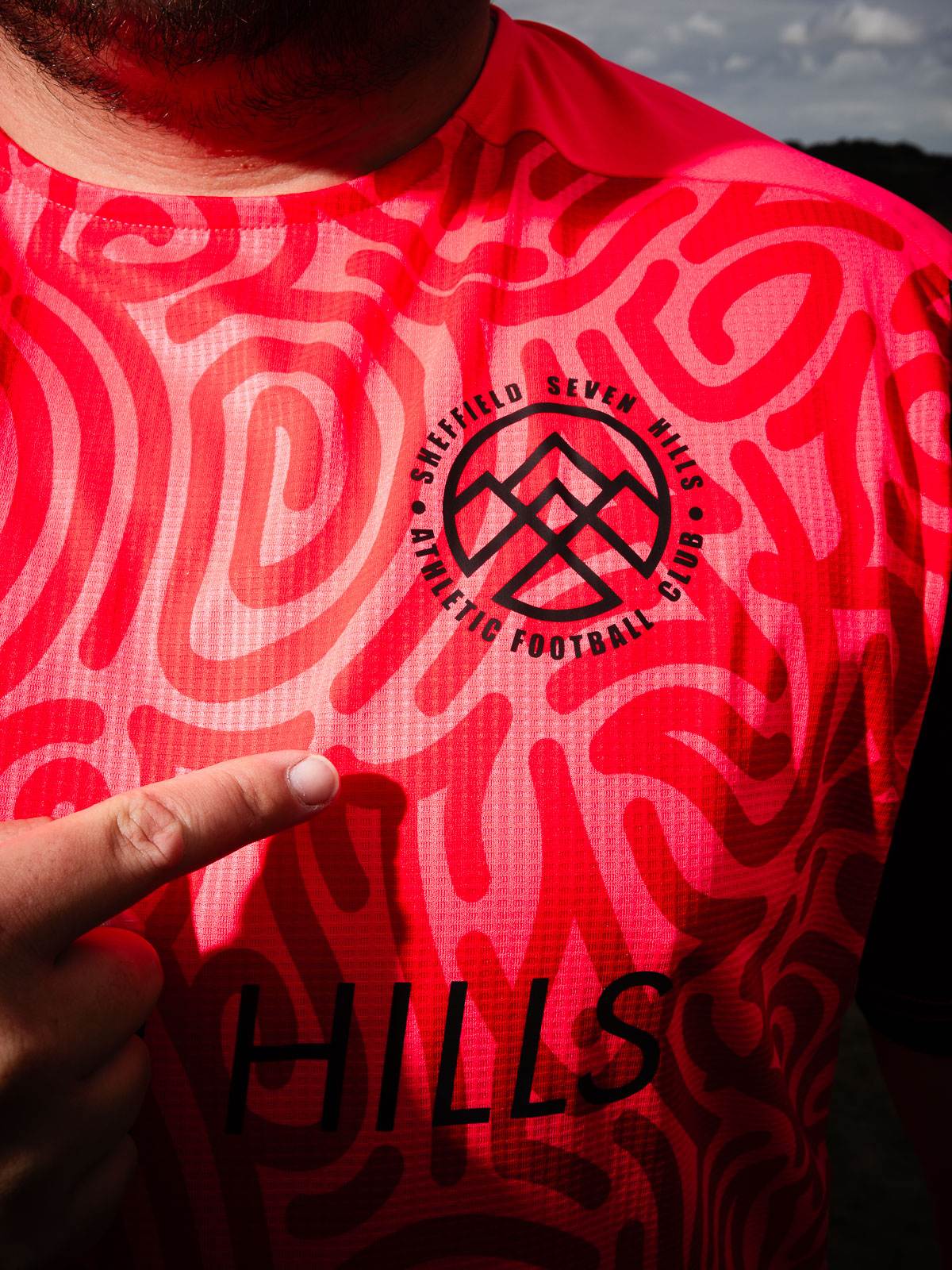 Player wearing new pink Sheffield Seven Hills AFC away kit pointing ot the club badge.